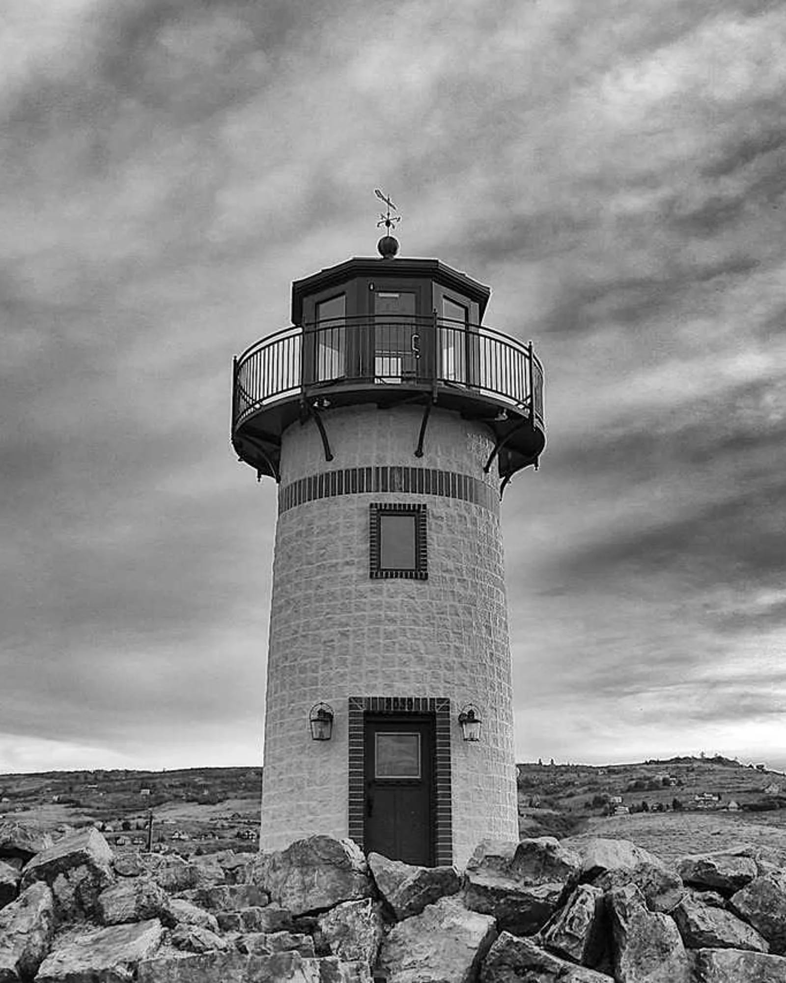 A black-and-white photograph of a small lighthouse on rocks under dramatic streaked clouds.