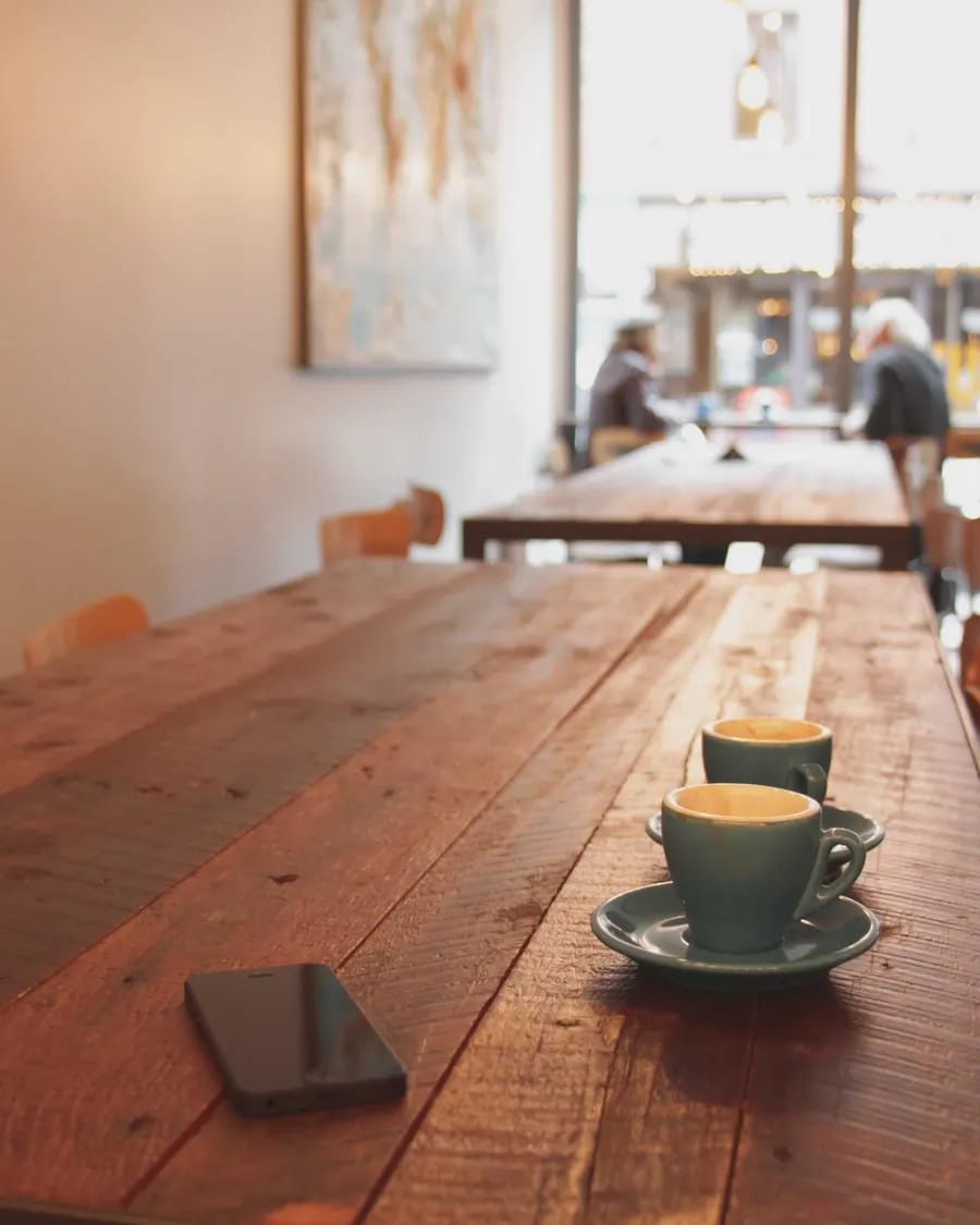 A long wooden table beside tall windows in a café, a coffee cup catching warm afternoon light.